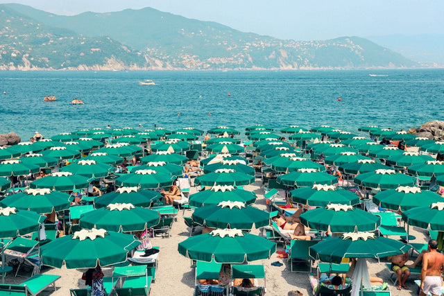 Colorful sunbeds and green parasols at an Italian beach print with blue water and hills in the background.